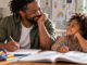 Ein Vater und sein Grundschulkind sitzen entspannt am Küchentisch und lächeln sich an. Vor ihnen liegen ein Schulheft, ein aufgeschlagenes Schulbuch und eine Federmappe.