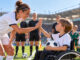 Eine Fußballkapitänin beugt sich auf Augenhöhe zu einem Jungen im Rollstuhl und gibt ihm ein High-Five. Im Hintergrund stehen Spieler*nnen, Schiedsrichter*innen und Einlaufkinder auf dem Fußballplatz.