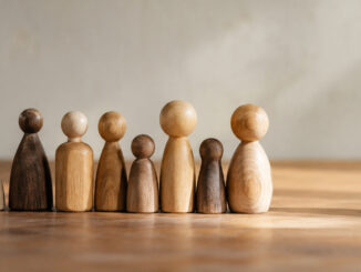 Eine Gruppe unterschiedlich großer und geformter Holzfiguren steht in einer Reihe. Eine einzelne Holzfigur steht mit etwas Abstand daneben. KI-generiertes Bild.
