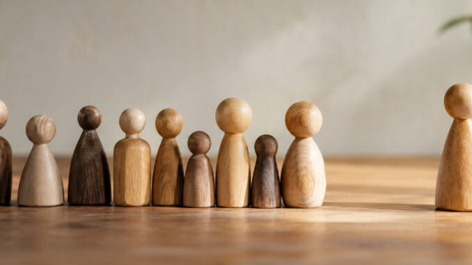 Eine Gruppe unterschiedlich großer und geformter Holzfiguren steht in einer Reihe. Eine einzelne Holzfigur steht mit etwas Abstand daneben. KI-generiertes Bild.