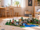 Helles, aufgeräumtes Kinderzimmer mit Spielzeugregalen, Bett und Schreibtisch. Auf dem Teppich steht eine große, aufgebaute Landschaft aus bunten Klemmbausteinen.